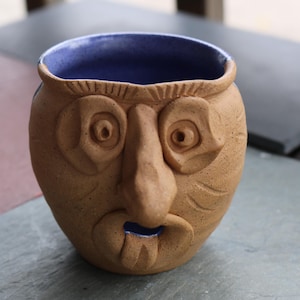 May include: A handmade ceramic mug with a face design. The mug is tan with a blue interior and features carved facial details including eyes, a nose, and a mouth. The mug is a unique piece of pottery.