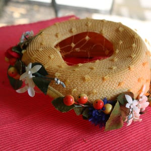 May include: A vintage yellow straw hat with a circular design and a decorative floral arrangement. The hat features a dotted pattern and is adorned with red berries, white flowers, and green leaves. A delicate net veil is visible inside the hat.