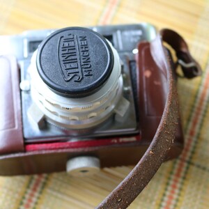 Pu&ograve; includere: Una custodia per macchina fotografica vintage in pelle marrone con una macchina fotografica argentata e nera all'interno. L'obiettivo della fotocamera ha un tappo nero con la scritta "STEINHEIL MUNCHEN" stampata su di esso.