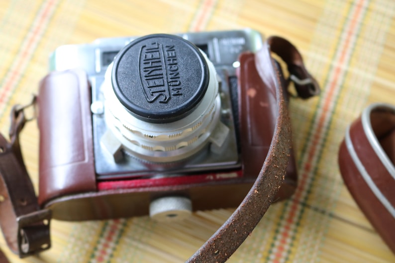 Pu&ograve; includere: Una custodia per macchina fotografica vintage in pelle marrone con una macchina fotografica argentata e nera all'interno. L'obiettivo della fotocamera ha un tappo nero con la scritta "STEINHEIL MUNCHEN" stampata su di esso.