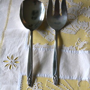 May include: A silver serving spoon and fork set. The spoon has a large, oval bowl, and the fork has three tines. Both utensils have long, slender handles with a decorative floral design at the base. The set is displayed on a white and yellow lace tablecloth.