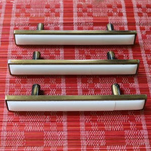 Könnte beinhalten: Drei Schrankgriffe mit einem weißen rechteckigen Mittelteil, umrahmt von goldfarbenem Metall. Jeder Griff hat zwei dunkle Metallpfosten. Die Griffe sind auf einer roten gewebten Oberfläche angeordnet.