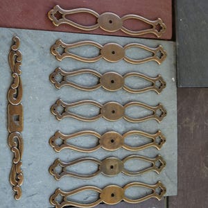 May include: A collection of seven elongated brass drawer pulls with an openwork design and a central circular mounting hole. One additional vertical brass hardware piece features a central square opening and decorative scrollwork at both ends.