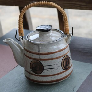 May include: A ceramic teapot with a woven handle and lid. The teapot is off-white with brown stripes and circular designs. The handle is made of woven light brown material. The teapot is on a multi-colored surface.