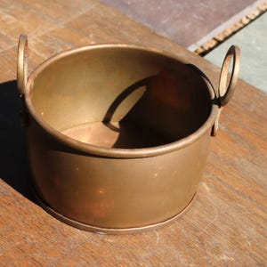 May include: A small, round, antique brass pot with two loop handles. The pot has a slightly flared rim and a simple, unadorned design. The brass has a warm, aged patina, suggesting it is vintage. The pot sits on a wooden surface.