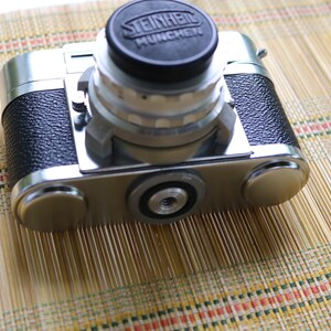 Pu&ograve; includere: Una macchina fotografica a pellicola da 35 mm argentata e nera con un obiettivo Steinheil Munchen. La macchina fotografica &egrave; appoggiata su un tappetino di bamb&ugrave; intrecciato.