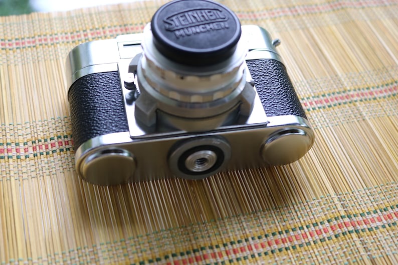 Pu&ograve; includere: Una macchina fotografica a pellicola da 35 mm argentata e nera con un obiettivo Steinheil Munchen. La macchina fotografica &egrave; appoggiata su un tappetino di bamb&ugrave; intrecciato.