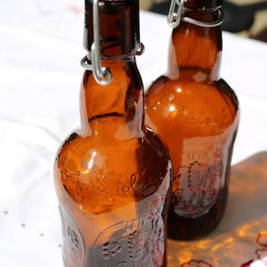 Peut inclure: Deux bouteilles en verre brun avec des bouchons à bascule. Les bouteilles portent une étiquette avec le mot "Grolsch" dessus.