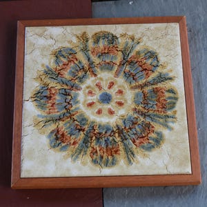 May include: A square ceramic tile framed in medium-toned wood featuring a central circular floral motif. The design displays a crackled texture with a color palette of muted blue, burnt orange, and cream, creating a weathered appearance.