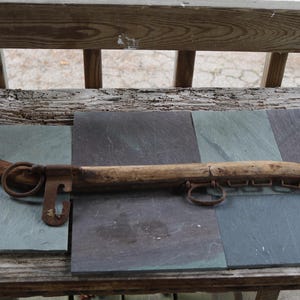 Op de afbeelding: Een antiek houten juk met metalen ringen en beugels. Het juk heeft een lichtbruine kleur en lijkt verweerd, wat duidt op leeftijd en gebruik. Het rust op een oppervlak van grijze en blauwe leistenen tegels, met een houten leuning op de achtergrond.