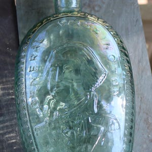 May include: A vintage, oval-shaped, light green glass bottle. The bottle features an embossed portrait of a person and the words "GENERAL WASHINGTON" around the edge. The bottle has a rounded base and a narrow neck.