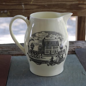 May include: Cream-coloured ceramic jug with a black and white transferware design. The design shows a historical scene with a building, figures, and trees. The jug has a curved handle and a spout for pouring.