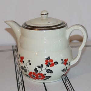 Puede incluir: Tetera de cerámica color crema con tapa y asa. La tetera presenta un borde marrón y un diseño floral con flores rojas y hojas negras. La tetera está diseñada para preparar té.