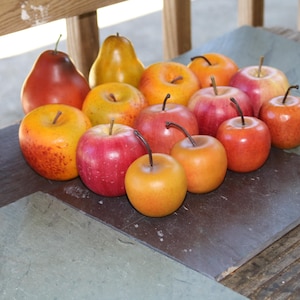 May include: A collection of artificial fruit including several red, yellow, and orange apples and two pears. Each piece of fruit features a smooth, rounded surface and a small, dark brown stem protruding from the top, arranged in a cluster.