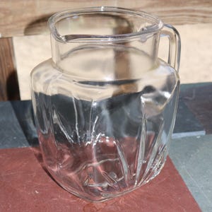May include: A clear glass pitcher with a handle and a spout. The pitcher has a geometric design on the sides and is sitting on a red surface. The background is a mix of wood and other surfaces.