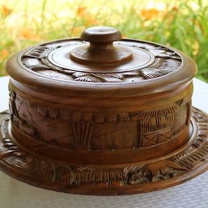 May include: A round wooden box with a lid, intricately carved with a scene of houses and trees. The box is brown and has a smooth, polished finish.