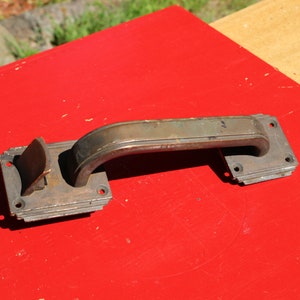 May include: A vintage bronze door handle with a curved design and a latch mechanism. The handle is mounted on a red surface.