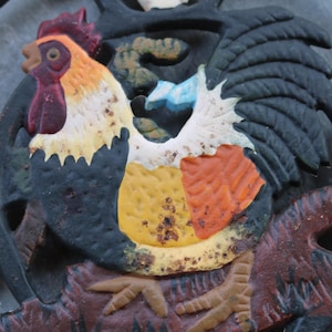 May include: A cast iron rooster design with a colorful, detailed depiction of a rooster. The rooster has a red comb, yellow and orange body, and black tail feathers. The design includes floral accents and is set against a dark background.