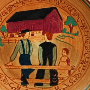 May include: A ceramic plate with a brown background and a green border. The center of the plate features a painted scene of three people in front of a red barn. The people are wearing traditional clothing and are sitting on a bench. The plate is decorated with a brown and green floral pattern around the edge.