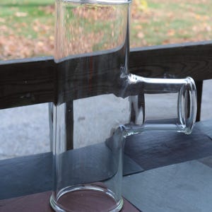 May include: Clear glass T-shaped bong with a cylindrical chamber and a horizontal mouthpiece. The bong is made of transparent glass, allowing visibility of the interior. The design features rounded edges and a sturdy base.