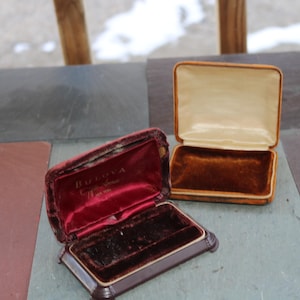 Puede incluir: Dos estuches de reloj Bulova vintage. Un estuche está abierto, revelando un interior de terciopelo rojo con el logotipo de Bulova. El otro estuche está abierto, mostrando un interior de terciopelo marrón. Ambos estuches son marrones con ribetes dorados.