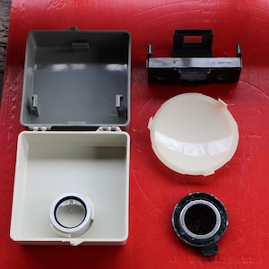 May include: An assortment of vintage camera accessories, including a gray and cream-colored case, a black and silver lens, a black viewfinder, and a cream-colored lens cover. The items are arranged on a red surface.