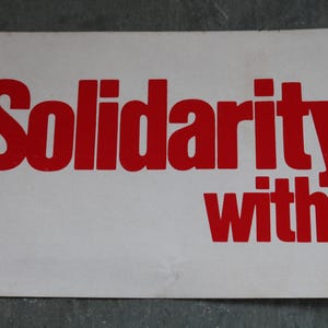 Può includere: Un cartello rettangolare bianco con testo rosso in grassetto che recita Solidarity with. Le lettere sono stampate in uno stile sans-serif, con la parola Solidarity posizionata sopra la parola with su uno sfondo bianco uniforme.