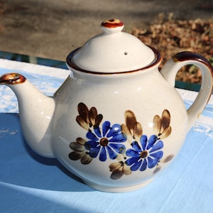 May include: A white ceramic teapot with a brown lid and handle. The teapot is decorated with brown leaves and blue flowers.