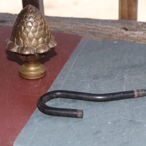 Może przedstawiać: Mosiężny, dekoracyjny przedmiot w kształcie żołędzia o szczegółowej, teksturowanej powierzchni, obok zakrzywionego, czarnego metalowego pręta z gwintowanymi końcami. Przedmioty znajdują się na dwutonowej powierzchni w kolorze czerwonym i szarym.