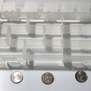 May include: A clear plastic storage box with multiple compartments. The box is empty and has a rectangular shape. Three US coins are visible in the foreground.