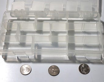 Op de afbeelding: Een transparante plastic opbergdoos met meerdere vakken. De doos is leeg en heeft een rechthoekige vorm. Drie Nederlandse munten zijn zichtbaar op de voorgrond.