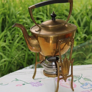 Könnte beinhalten: Eine Messing-Teekanne mit schwarzem Griff steht auf einem Ständer mit einem Brenner. Die Teekanne hat einen perlenbesetzten Rand und einen dekorativen Knauf auf dem Deckel. Der Ständer hat drei Beine und ist mit einem floralen Muster verziert.