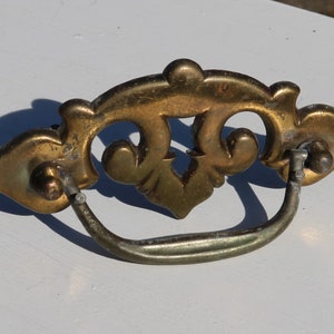 Op de afbeelding: Een messing ladegreep met een decoratief, sierlijk ontwerp. De greep heeft een lusvormige handgreep en twee schroefgaten.