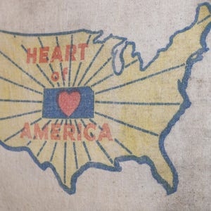 May include: A beige fabric banner featuring a stylized map of the United States. The map is outlined in blue, with a yellow interior and radiating lines. The words "HEART of AMERICA" are in red, with a red heart over a blue rectangle.