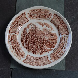 以下が含まれることがあります： 白のセラミックプレートに茶色の転写デザイン。プレートには、船と人物が登場する航海のシーンが描かれ、装飾的なボーダーとテキストで囲まれています。プレートはスカラップエッジです。