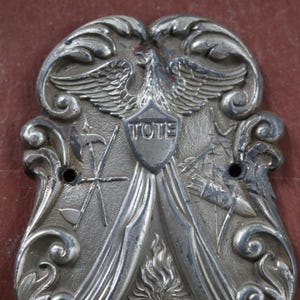 May include: Ornate silver-toned metal emblem with an eagle, shield, and the word "TOTE." The design includes crossed tools, flames, and the letters "E. F. C." Decorative scrollwork frames the central elements. Two holes are visible.