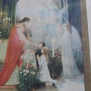 May include: A vintage religious print depicting a priest in a red robe giving communion to a young girl. The girl is kneeling and receiving the host from the priest. A white-robed angel stands behind the girl, with a halo above her head. The scene is set in a church with a floral arrangement in the foreground.