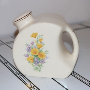 May include: Cream-coloured ceramic pitcher with a floral design featuring yellow and purple flowers. The pitcher has a handle and a spout with a lid. Gold trim accents the lid and handle. The pitcher is likely vintage.