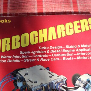 Puede incluir: Un libro rojo titulado "Turbochargers" con un título amarillo. La portada del libro presenta un primer plano de un turbocompresor plateado con mangueras azules. El texto de la portada del libro describe el contenido del libro, incluyendo el diseño del turbocompresor, el dimensionamiento, la adaptación, el encendido por chispa, las aplicaciones de motores diésel, la inyección de agua, los controles, la carburación, la refrigeración intermedia, los detalles de instalación, los coches de calle y de carreras, los barcos y las motocicletas.
