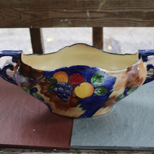 May include: A vintage ceramic serving bowl with a cream-colored interior and a scalloped rim. The exterior features a hand-painted design of fruit, including grapes, oranges, and apples, set against a blue and brown background. The bowl has two dark blue handles.