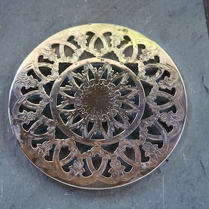 Puede incluir: Salvamanteles plateado ornamentado con un diseño circular. El salvamanteles presenta un patrón central de rayos de sol rodeado de intrincados calados con formas florales y geométricas. El salvamanteles de metal está diseñado para proteger las superficies.