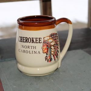 Pode incluir: Caneca de cerâmica creme com borda e alça marrons. A caneca apresenta o texto "CHEROKEE NORTH CAROLINA" e uma ilustração de um nativo americano com cocar. A caneca está sobre uma superfície cor de ardósia.