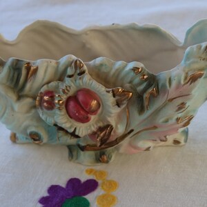 May include: A vintage ceramic planter with a leaf design and gold accents. The planter is painted in shades of blue, pink, and green, and features a raised floral design with red accents.