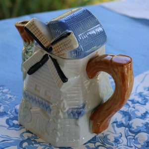 May include: A white ceramic teapot shaped like a windmill with blue and brown accents. The teapot has a spout and a handle.