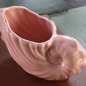 Może przedstawiać: Jasnoróżowa ceramiczna doniczka w kształcie muszli. Naczynie ma błyszczące wykończenie z głębokimi, pionowymi żłobieniami, które podążają za naturalną spiralną krzywizną muszli, tworząc u góry wydrążony otwór na drobne przedmioty.