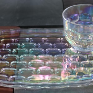 Puede incluir: Juego de taza y bandeja de té de vidrio iridiscente. El vidrio transparente tiene un patrón texturizado y un brillo de arcoíris. La bandeja tiene asas y un borde festoneado. La taza se asienta sobre una sección de bandeja más pequeña a juego.