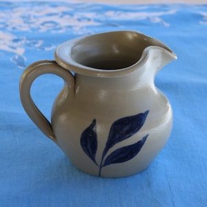 Könnte beinhalten: Ein kleiner, grauer Keramik-Krug mit einem blauen Blatt-Design. Der Krug hat einen runden Körper und einen Henkel.