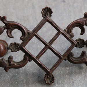 Könnte beinhalten: Verziertes, dunkelbraunes Metallmöbelbeschlag. Das Stück hat ein zentrales rautenförmiges Gitterdesign, flankiert von sich schlängelnden floralen und blattartigen Elementen sowie stilisierten Tiergesichtern. Wahrscheinlich für eine Schublade oder einen Schrank.