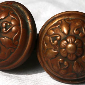 May include: Two antique copper door knobs with floral and leaf designs. The knobs are round with a raised floral pattern in the center, surrounded by leaves and other decorative elements. The copper has a warm, aged patina.