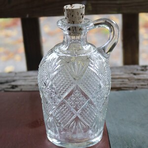 May include: Clear glass decanter with a diamond pattern and a handle. The decanter has a cork stopper. The handle is arched and attached to the neck of the decanter. The decanter is likely used for serving beverages.